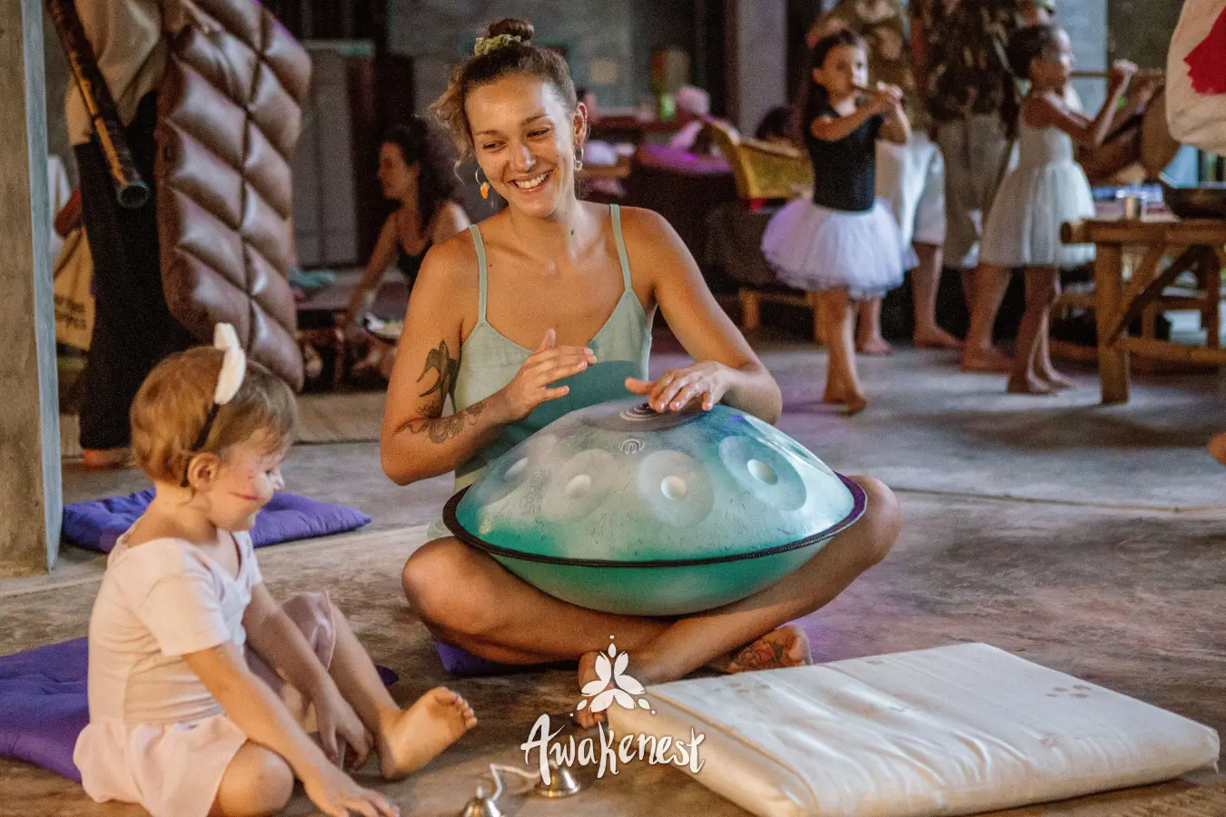 A woman plays a handpan beside a child seated on a mat in an open pavilion.