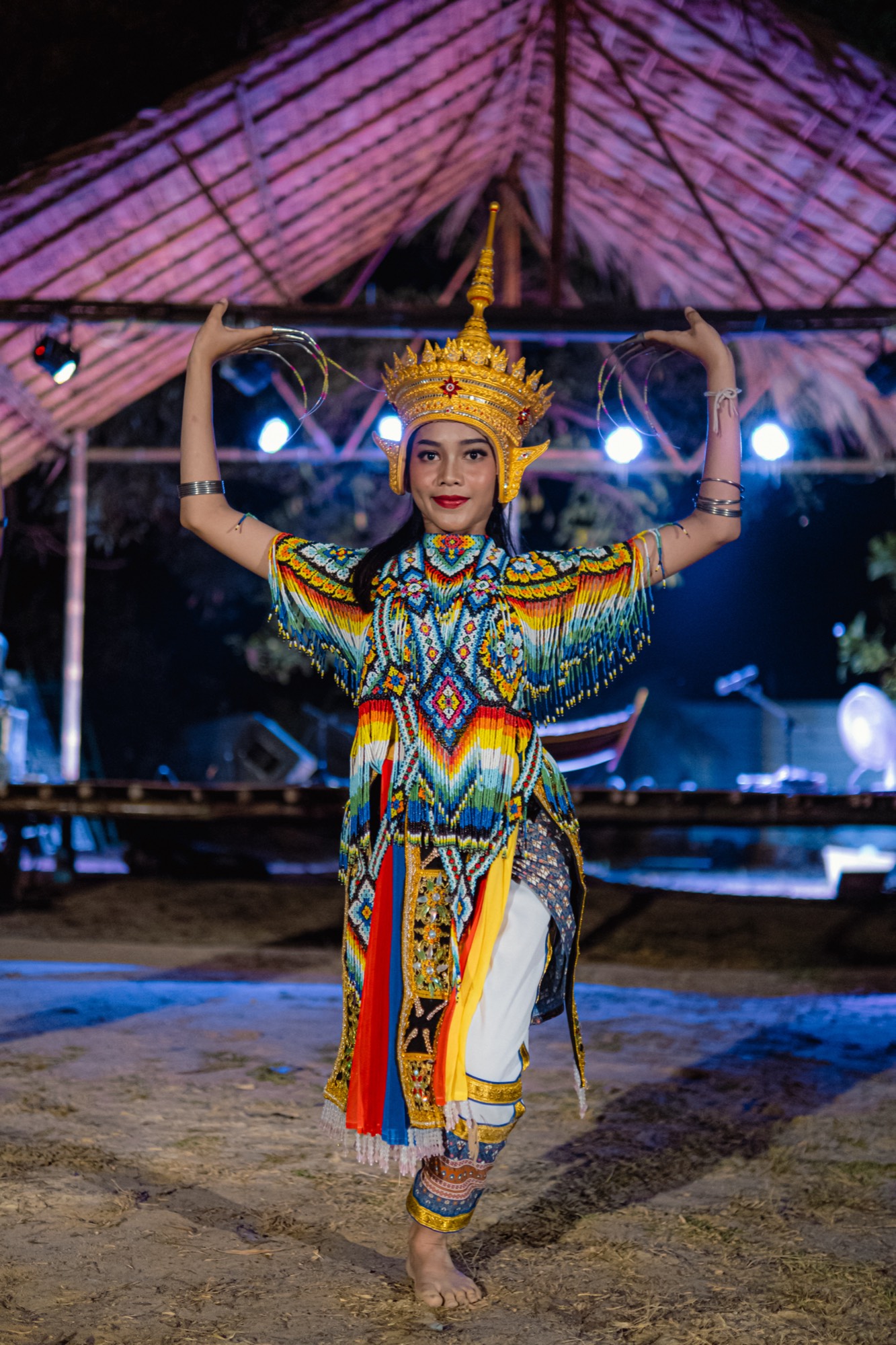 A dancer in a beaded costume and gold crown raises both arms in a classical pose on sand at night.
