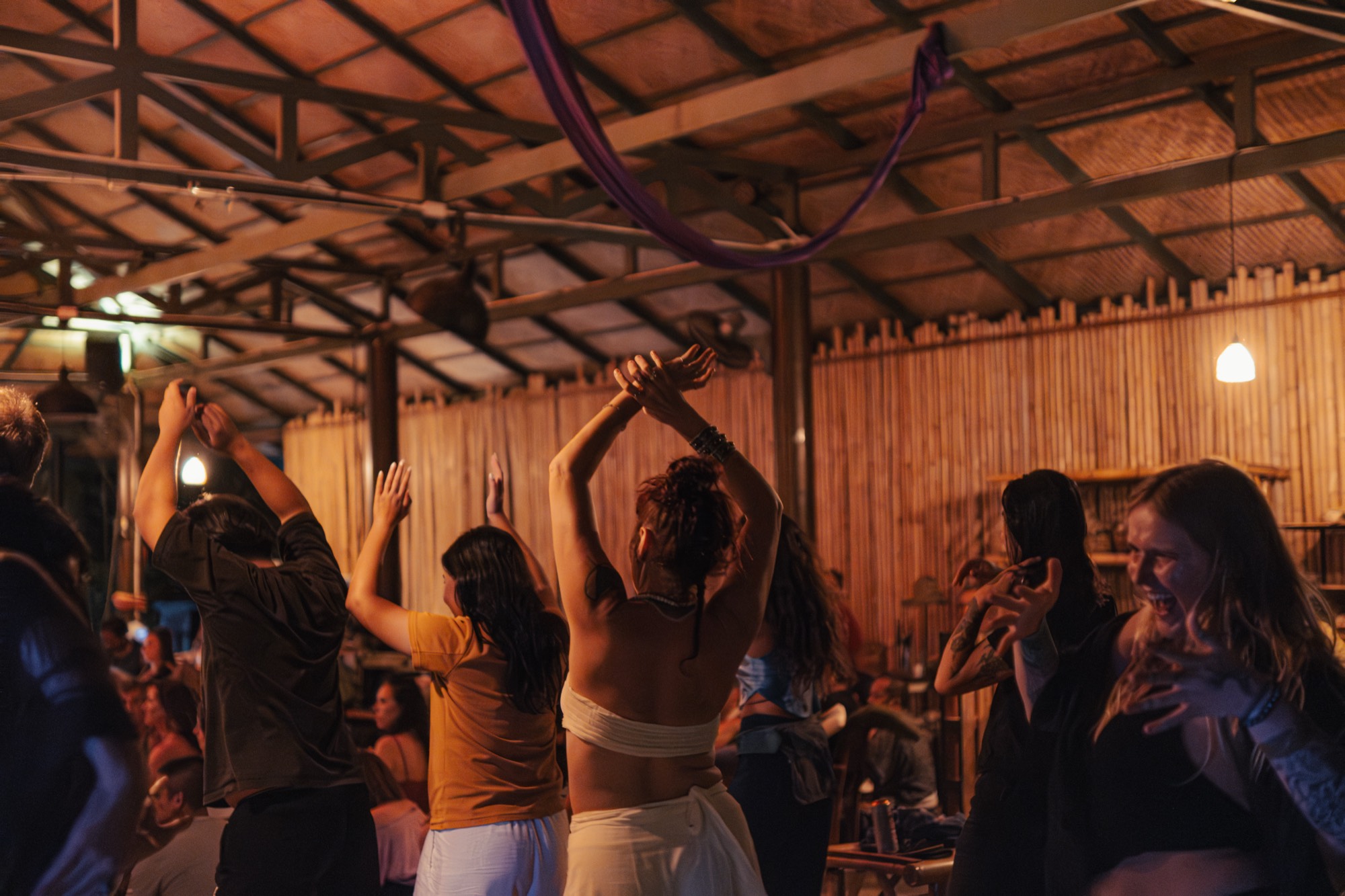 People dance with their arms raised under a timber and bamboo roof lit by warm lamps at night.