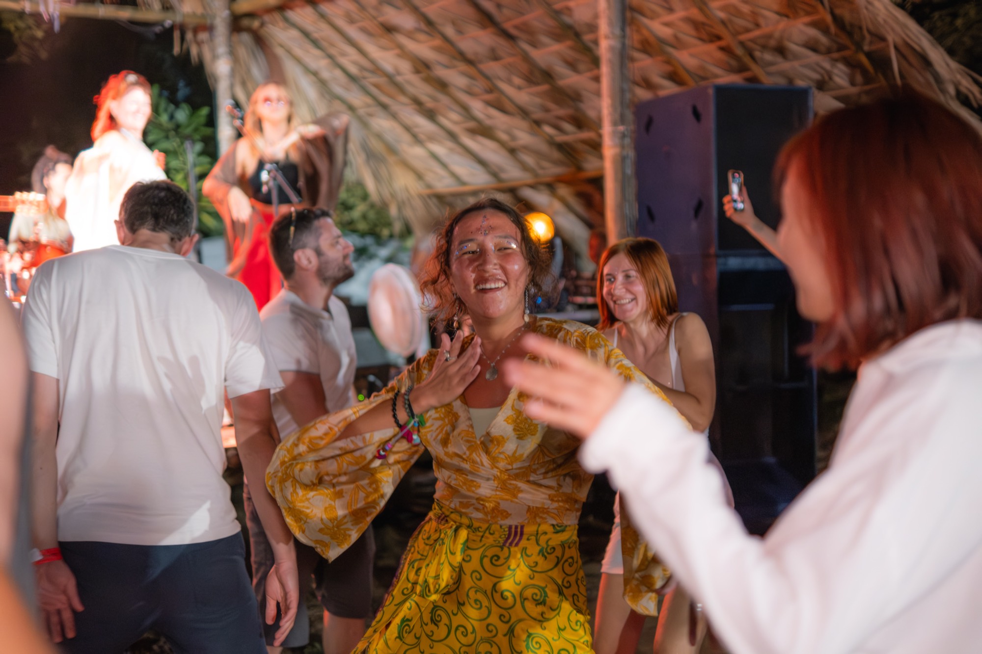 A woman in a yellow patterned dress laughs while dancing among other people under a thatched roof at night.