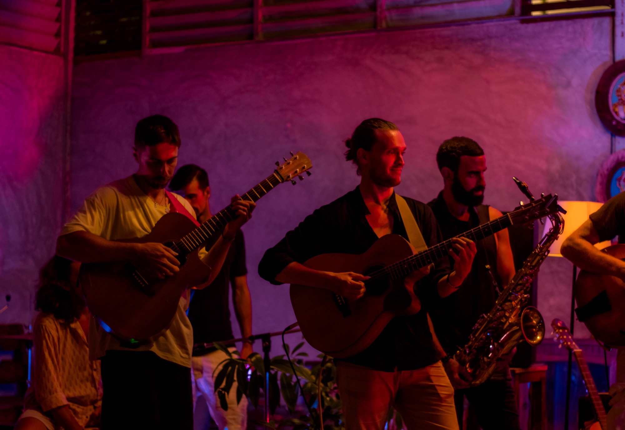 Four musicians play acoustic guitars and a saxophone side by side against a plaster wall under coloured light.